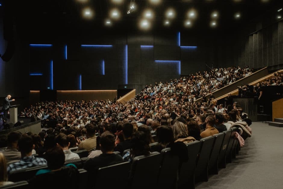 Fullsatt konferansesal sett bakfra med taler på opplyst scene